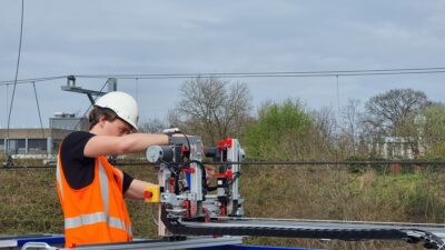 De studenten ontwikkelden een robot die het bevestigen van rijdraadklemmen automatiseert (hier wordt de robot klaargemaakt voor de demo, vlak voordat de zon gaat schijnen) Student maakt de robot klaar voor de demo