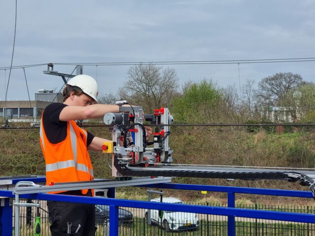 De studenten ontwikkelden een robot die het bevestigen van rijdraadklemmen automatiseert (hier wordt de robot klaargemaakt voor de demo, vlak voordat de zon gaat schijnen) Student maakt de robot klaar voor de demo